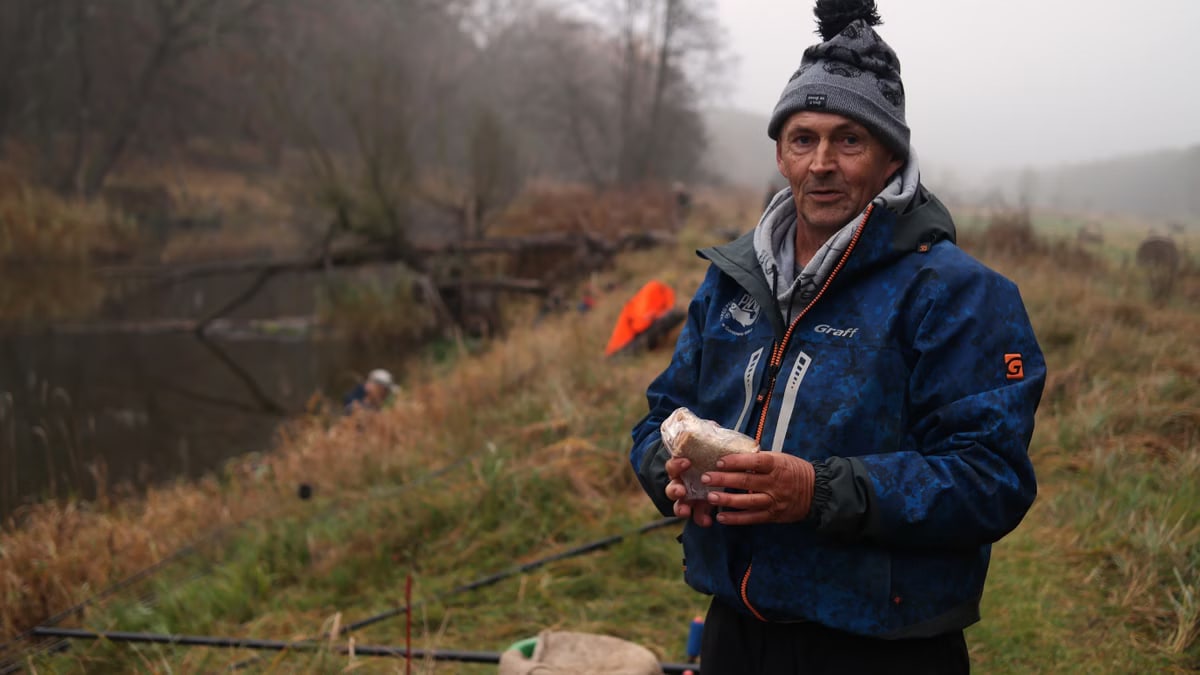 Man in warm clothing holding bait by a misty river, ready for fishing on a foggy day.